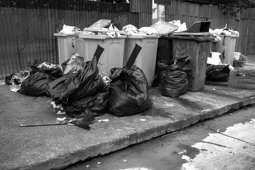 A row of four large wheelie bins positioned on a concrete pavement in an outdoor area near a wooden fence, with the two bins on the left being light grey and the two on the right in darker shades. The bins are filled with various waste materials, including flattened cardboard boxes, plastic packaging, and miscellaneous rubbish. Several black refuse bags are piled around the base of the bins, some leaning against them and others lying flat on the ground. The pavement shows signs of minor wear and dirt, and the environment appears typical of a residential or commercial rubbish collection point, consistent with waste removal services offered by Garden Clearance Belmont in the Belmont area. The scene is captured during daytime with natural light illuminating the scene uniformly, emphasizing the textures of the plastic bags, cardboard, and the surface of the bins. The overall setting reflects a typical rubbish collection or clearance situation in a suburban or urban locale, relevant to local waste management and rubbish removal services in the Belmont vicinity.