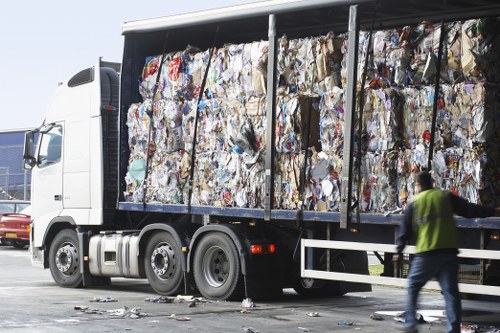 A large flatbed lorry parked on a paved surface is loaded with tightly compressed bales of mixed waste paper and cardboard materials, visible through a metal frame side panel. In the foreground, a person wearing a high-visibility vest is walking past the vehicle, which is part of a waste removal operation associated with rubbish collection and disposal services. The background shows a clear sky and other vehicles, suggesting a busy outdoor environment, possibly near a commercial or industrial site. The scene reflects typical rubbish transportation used by companies like Garden Clearance Belmont, serving areas including Belmont in the vicinity of London, with the lorry's load highlighting the storage of recyclable and general waste materials ready for collection or processing.