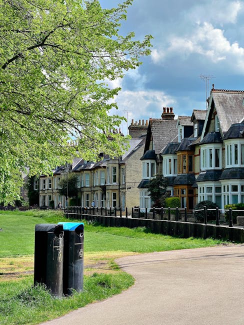 A peaceful residential area featuring a row of traditional Victorian-style terraced houses with bay windows and slate roofs, situated behind a green lawn and a black safety fence. In the foreground, t